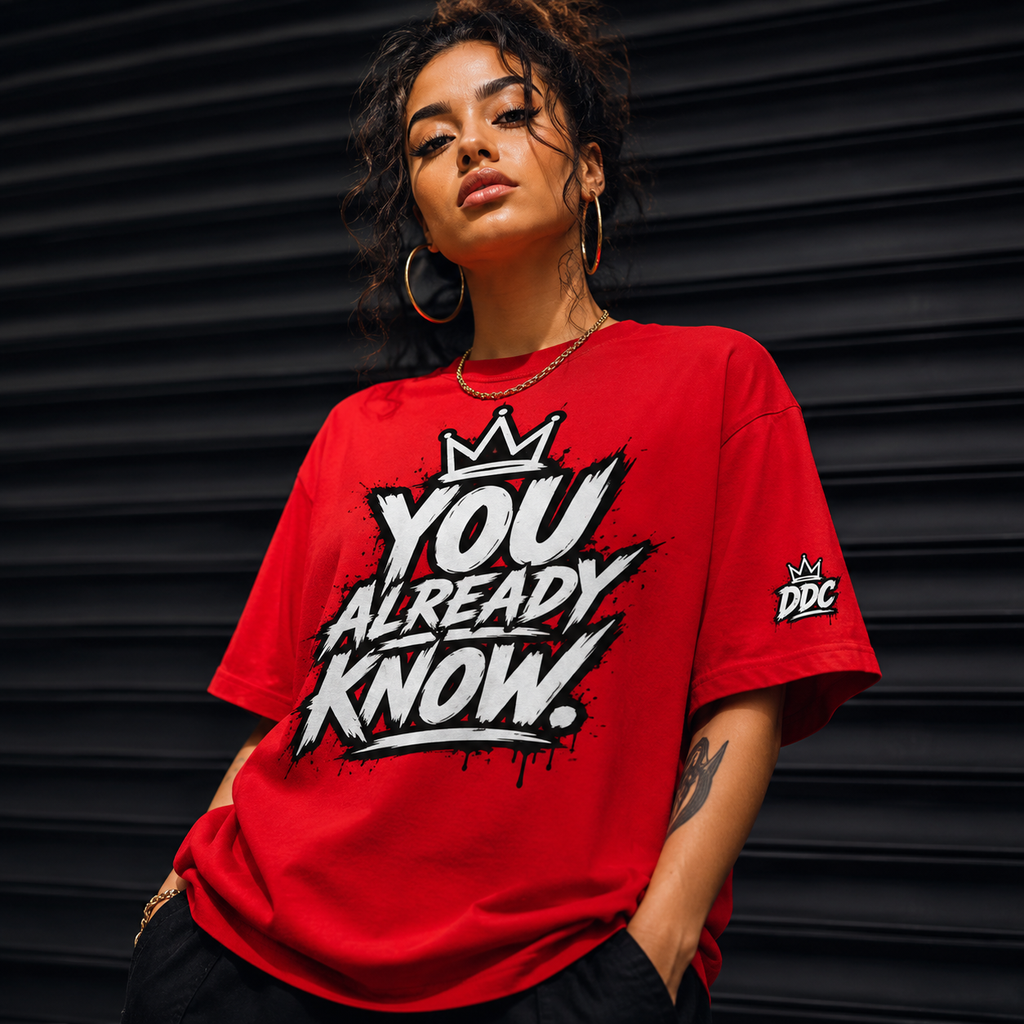 A woman wearing a bright red graphic t-shirt with the text 'YOU ALREADY KNOW.' in white and black graffiti-style lettering, featuring a crown logo on the chest and a 'DDC' logo on the sleeve, posed against a dark corrugated metal background.
