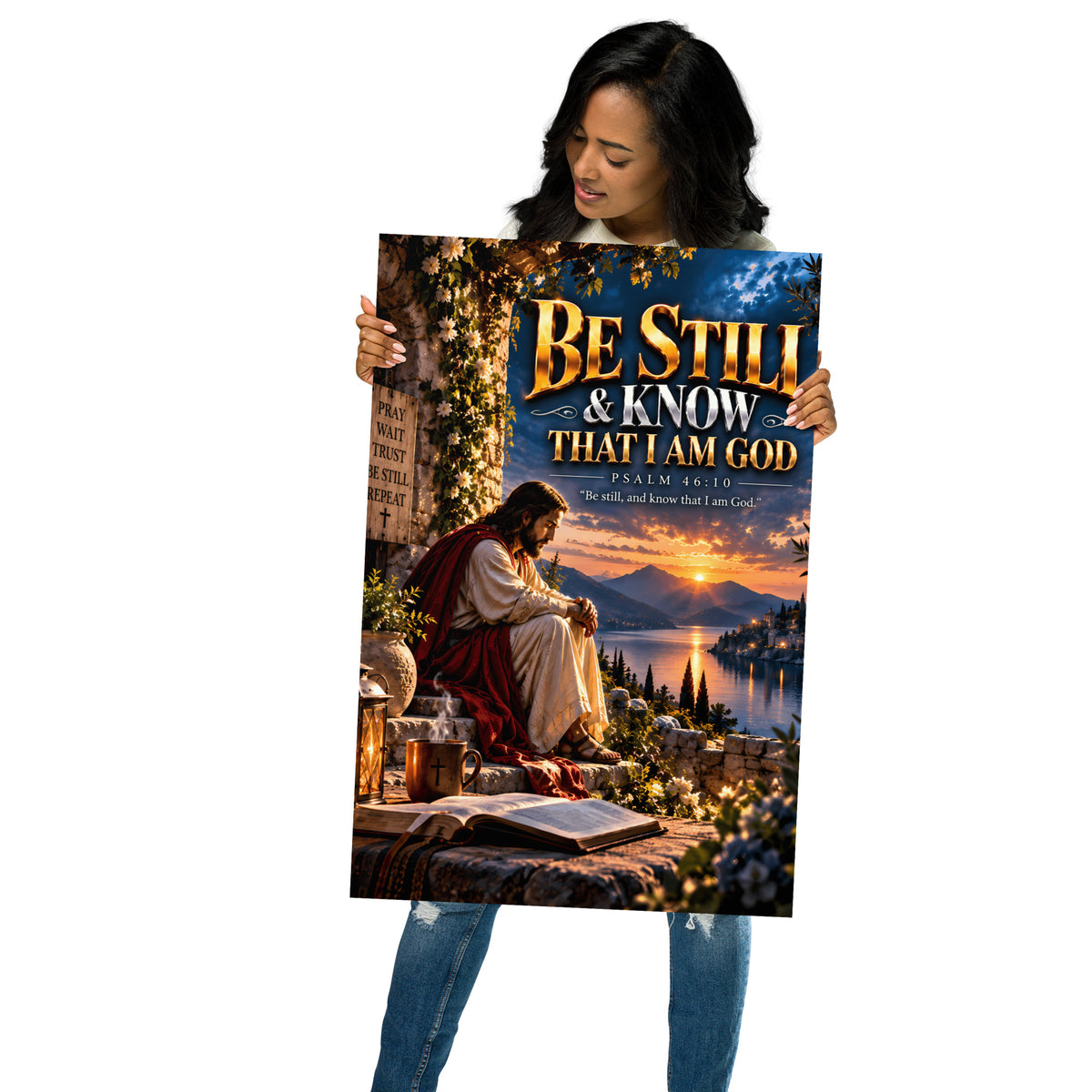 A woman holding a large vertical art print or poster featuring an illustrated scene of Jesus sitting by a lake at sunset, with the text 'BE STILL & KNOW THAT I AM GOD' and 'PSALM 48:10'. The background is plain white.