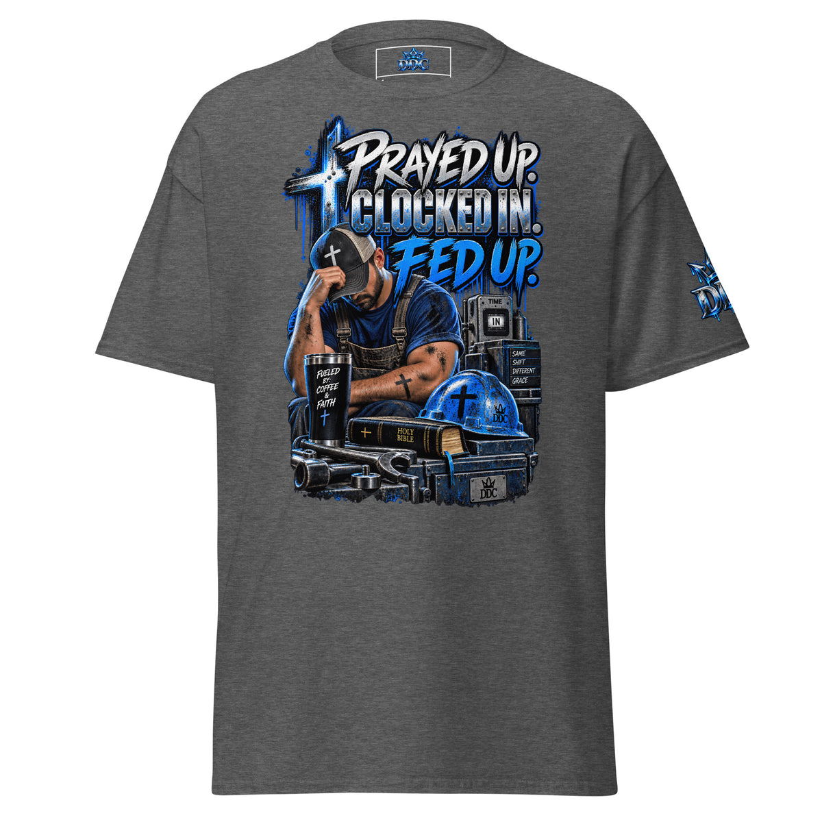 A dark heather gray t-shirt with a detailed graphic print on the front. The graphic features a man in a cap sitting at a desk with a cross on his head, surrounded by books, a mug, and a blue hard hat, with the text 'PRAYED UP.