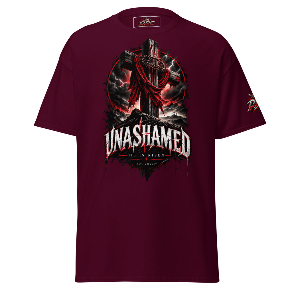 A maroon short-sleeve t-shirt with a large graphic print on the front featuring a cross, a draped figure, and the text 'UNASHAMED' and 'HE IS RISEN'.