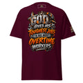 A maroon short-sleeve t-shirt with a large graphic on the front that reads 'GOD GIVES HIS TOUGHEST JOBS TO HIS MOST OVERTIME WORKERS' in orange, white, and metallic gray lettering, with a construction helmet and wrenches as part of the design. A smaller matching graphic is visible on the left sleeve.